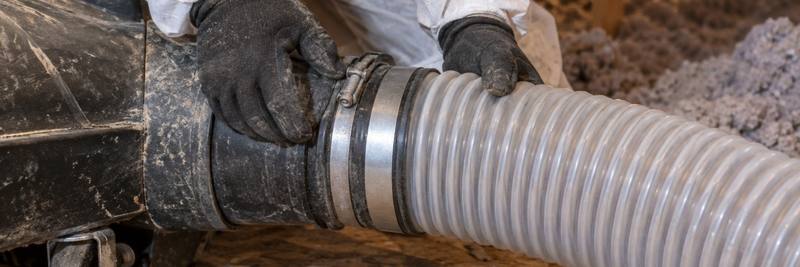 Gloved hands joining a grey insulation hose to a machine on an attic floor